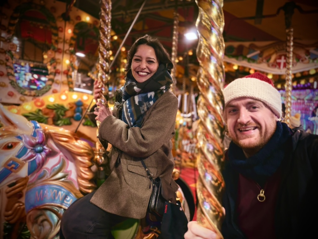 Marwa sitting on a carousel horse with Dan beside her, both wrapped up warm among the fairground lights.