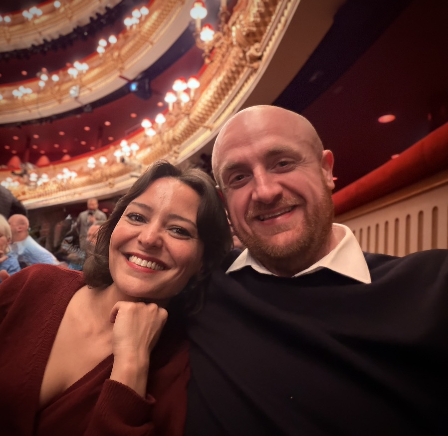 Marwa and Dan smiling together at the theatre, with the gilded balconies of the auditorium behind them.
